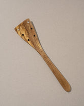 A wooden spatula with holes near the head, laid on a beige background.